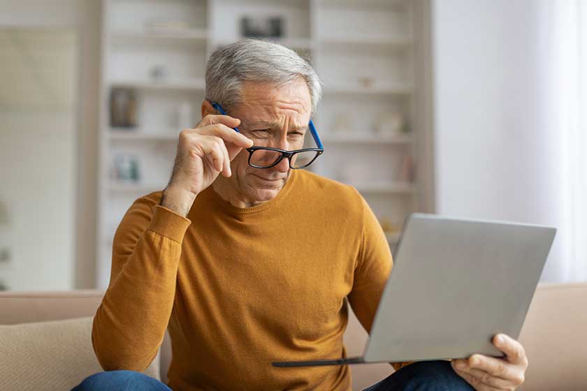 Focused elderly man making use of a laptop for either work or leisure at his residence, adjusting eyeglasses