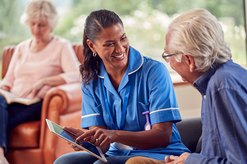 Senior Couple At Home With Man Talking To Female Nurse Or Care Worker Using Digital Tablet 